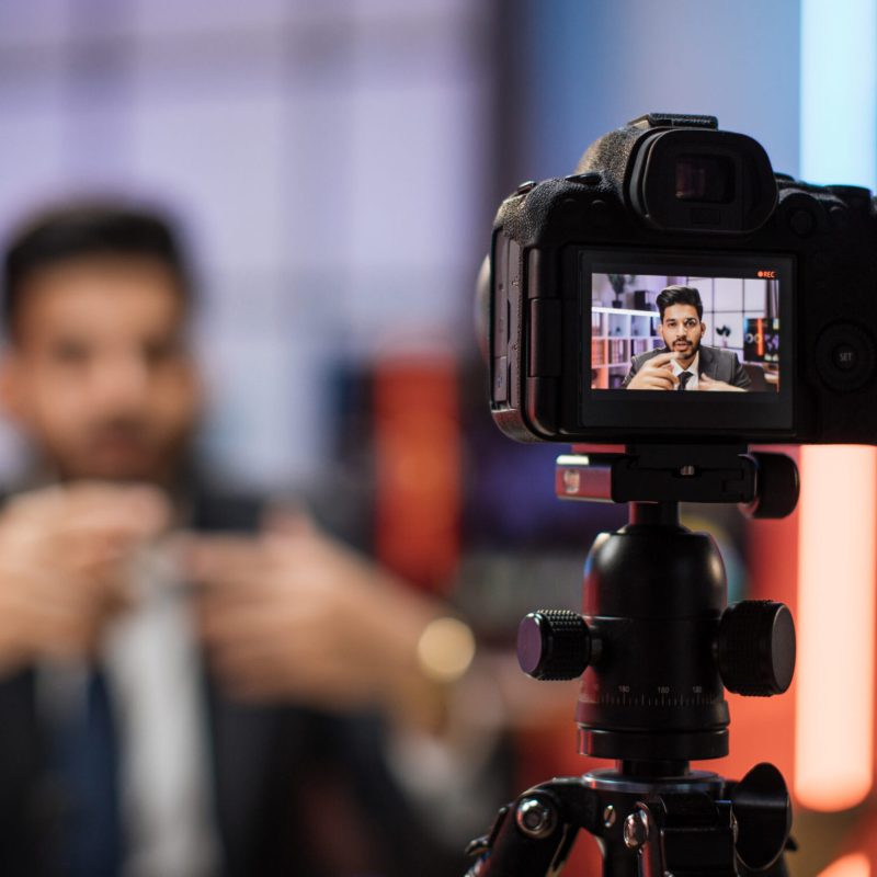 View from camera screen of confident indian bearded businessman, broker in suit sitting in front of camera in evening office during recording video for business vlog.